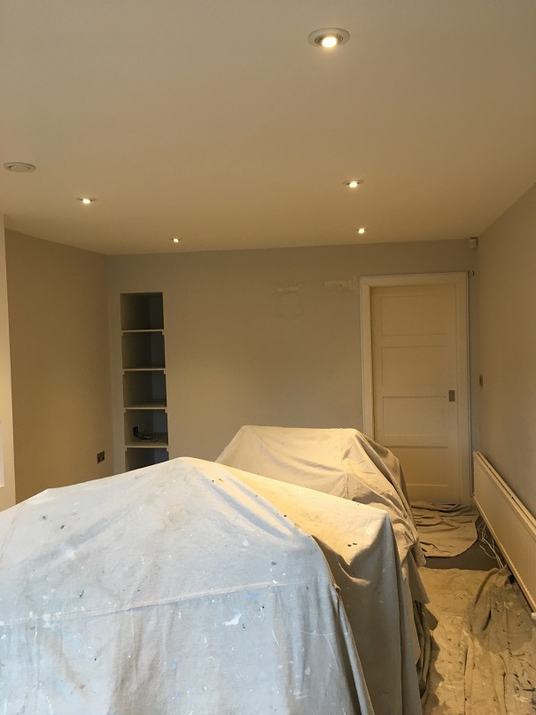 Before photo of grey room before Impressions Painters and Decorators wallpaper hanger re-decorated the room. Inbuilt shelves on left side of back wall. White door on right side of back wall. White radiator on right.side wall. Futniutre covered in dust sheets in centre of room