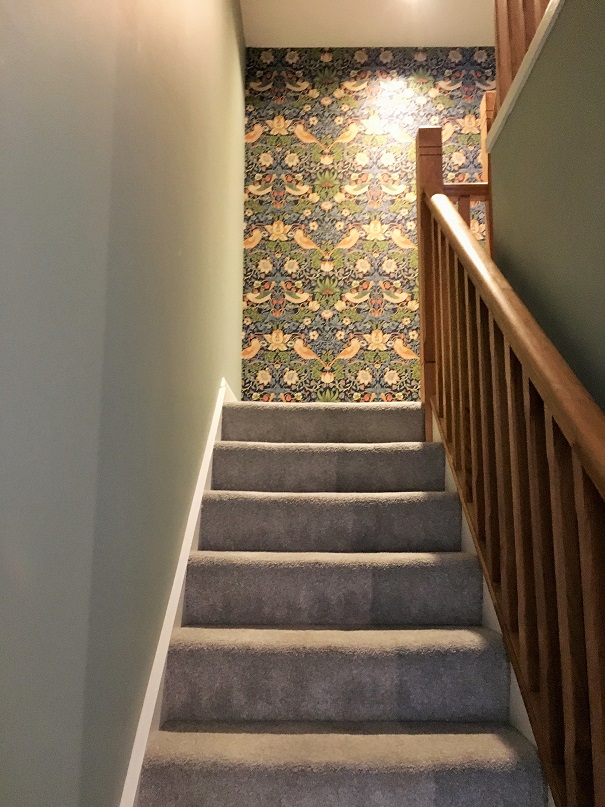 View looking up a flight of stairs to a turnabout on the stairs. Brown bannisters on right and green wall on left with grey carpet and white skirting. Impressions painters and decorators wallpapered wall the length of the wall on the turnabout. Pattered wallpaper with blue, green, orange, red and white with flowers leaves and birds.