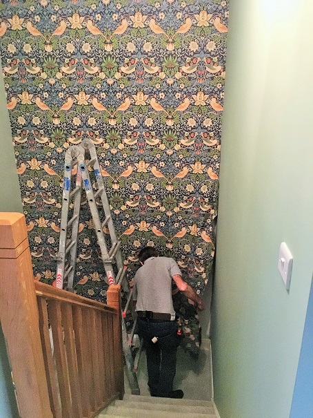 View looking down a flight of stairs to a turnabout on the stairs. Brown bannisters on left and green wall on right with a lightswitch. Impressions painters and decorators wallpaper hanger (man in blue jeans and grey t-shirt) hanging wallpaper the length of the wall on the turnabout with a ladder to his left. Pattered wallpaper with blue, green, orange, red and white with flowers leaves and birds.