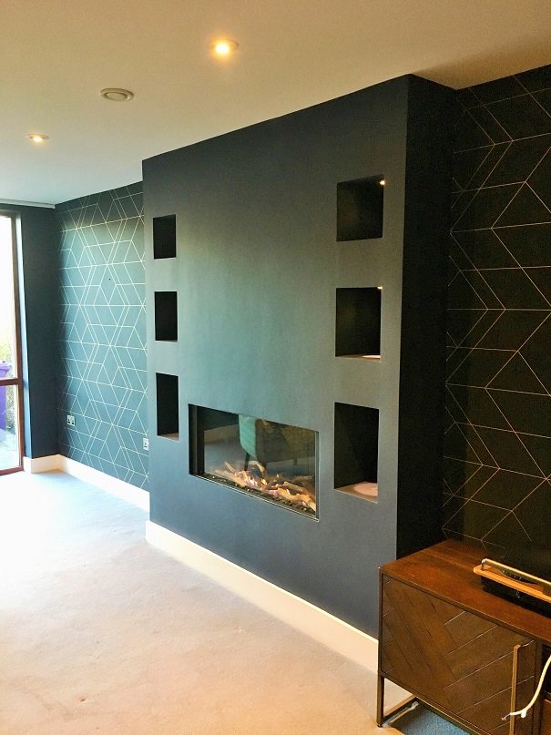 Right side view of a wallpapered wall by Impressions Painting and Decorating wallpaper hanger with a teal and white pattered wallpaper, and a centre wall painted teal with six alcoves on either side and a glass fronted fire at the bottom. Cream carpet with a partial view of a wooden sideboard on the front right