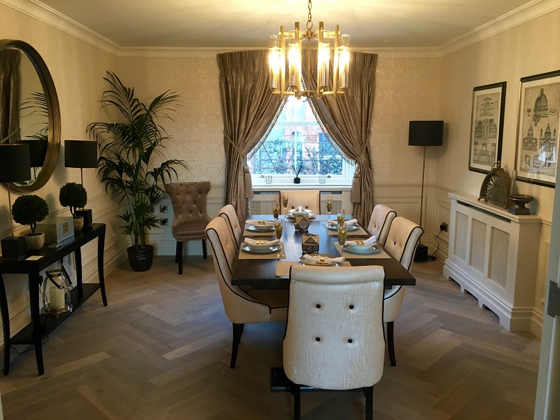 A dining room wallpapered with white pattered wallpaper by Impressions Painters and Decorators. From left to right; side table with round mirror above. Large plant in corner with blue velvet chair beside it, window with brown curtains, lamp in right corner, white radiator cover on right with two framed pictures of old architecture drawings above. Wood dining table with 6 white chairs with black legs and set for dinner in centre