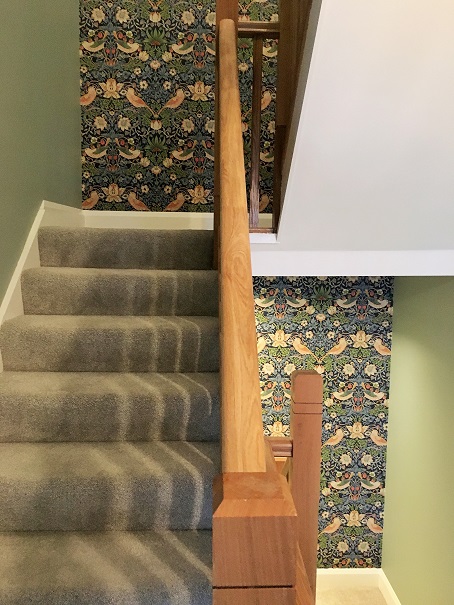 View looking up a flight of stairs to a turnabout on the stairs and down to next turnabout on left. Brown bannisters in centre and green walls on both sides. Pattered wallpaper with blue, green, orange, red and white with flowers leaves and birds on full length of walls on both turnabouts hung by Impressions painters and decorators wallpaper hanger