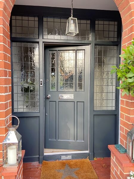A grey period door with glass panelling with small squares in the slightly opaque glass in the top half of the door and in the surrounds to the side set back behind the red brick surround of the house