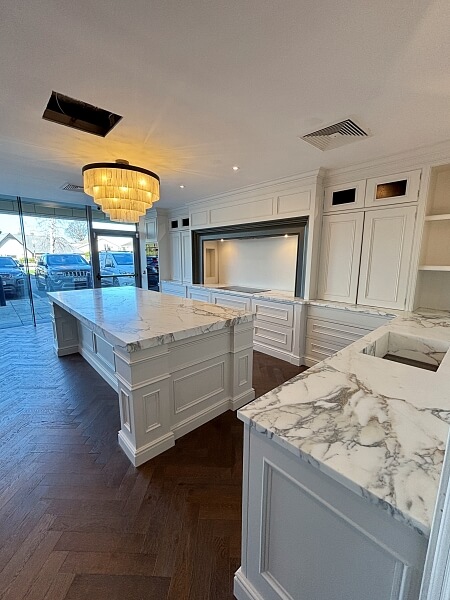 A white kitchen island with drawers and a white marble top with a chandelier in the centre and a wall in the background with white shelves and cabinets in the DeBarra Kitchen showroom after being hand painted by Impressions painting and decorating