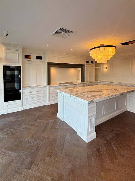 A white kitchen island with a white marble top in the centre right and a wall in the background with white shelves and cabinets, with a chandelier from the ceiling in the DeBarra Kitchen showroom after being hand painted by Impressions painting and decorating