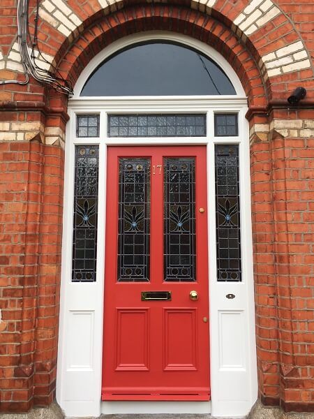 A red period door with stain glass panelling in the top half of the door and in the surrounds to the side and a clear fan glass panel on the top in a red brick surround of the house