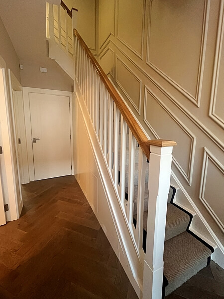 Side view of a white stairs and wood bannisters with full wall white stairs panelling in the background and a wooden floor after being painted by Impressions Painting and Decorating